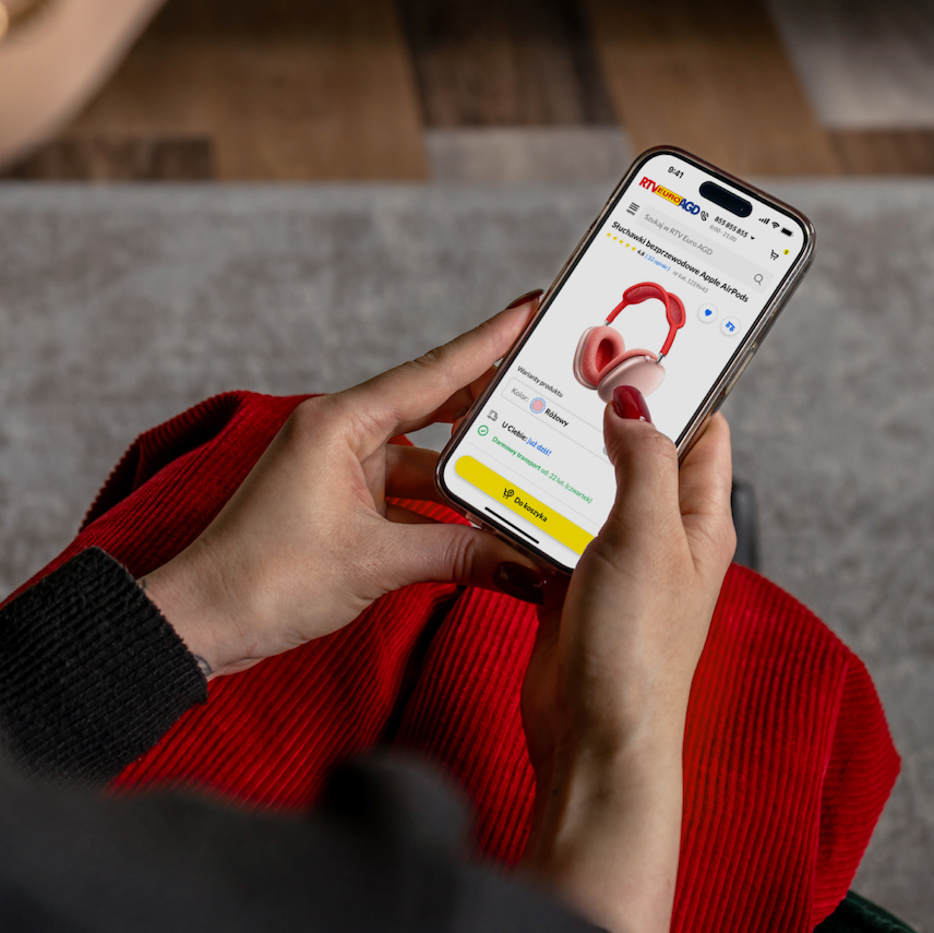 Zbliżenie na osobę trzymającą smartfon i korzystającą z aplikacji e-commerce RTV EURO AGD. Na ekranie widać różowe słuchawki Apple AirPods, wraz ze szczegółami produktu, opcjami wyboru koloru (