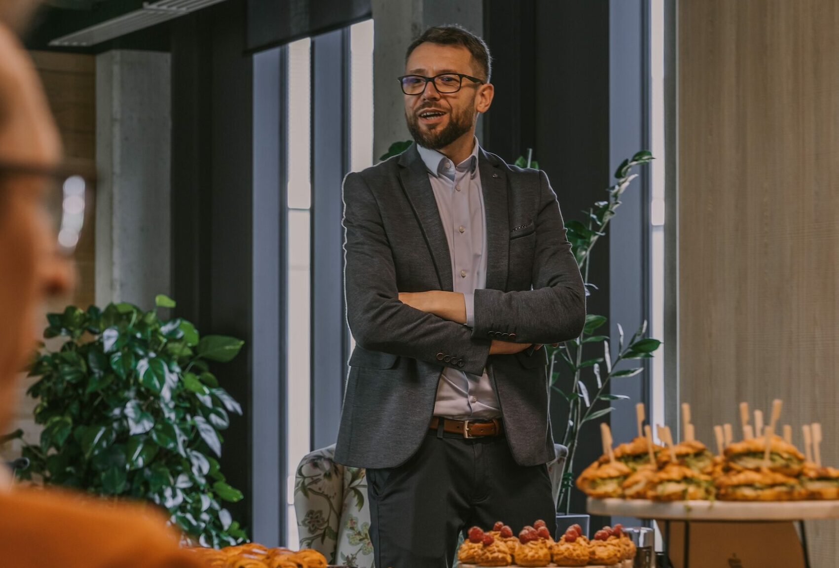 Grzegorz Jóźwiak - Biedronka - Mężczyzna w marynarce i okularach stoi przy stole i mówi do uczestników spotkania. Na pierwszym planie widać zastawiony stół z przekąskami i deserami. Spotkanie odbywa się w jasnym biurze z dużymi oknami i roślinami w tle.