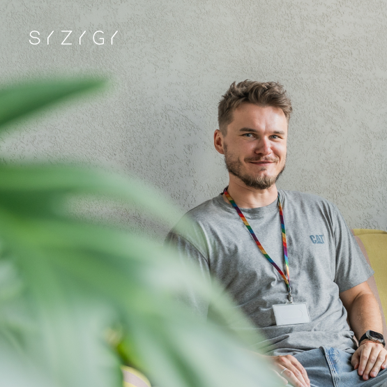 Marcin Stasiak, uśmiechnięty mężczyzna w szarej, casualowej koszulce, siedzący na żółtej kanapie w biurze SYZYGY Warsaw. Na szyi ma kolorowy smycz i identyfikator. Na pierwszym planie widoczna jest duża zielona roślina, dodająca naturalnego akcentu nowoczesnemu wnętrzu.