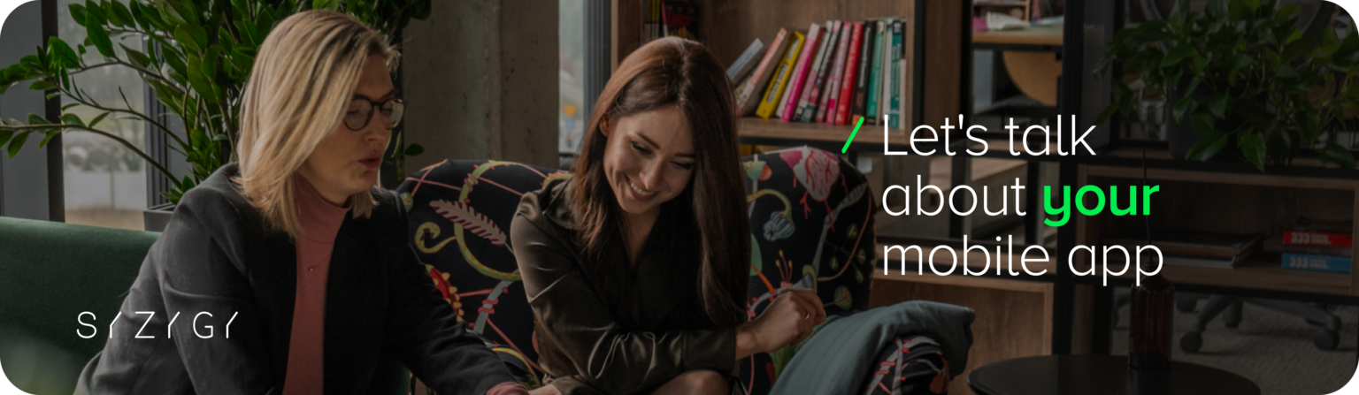 A photograph of two women sitting in a modern, cozy office space, engaged in a conversation and smiling while looking at a laptop. The background features bookshelves, plants, and a large window with natural light. Overlaid text reads, âLetâs talk about your mobile app,â emphasizing a collaborative and client-focused approach. The SYZYGY logo is placed in the bottom left corner.