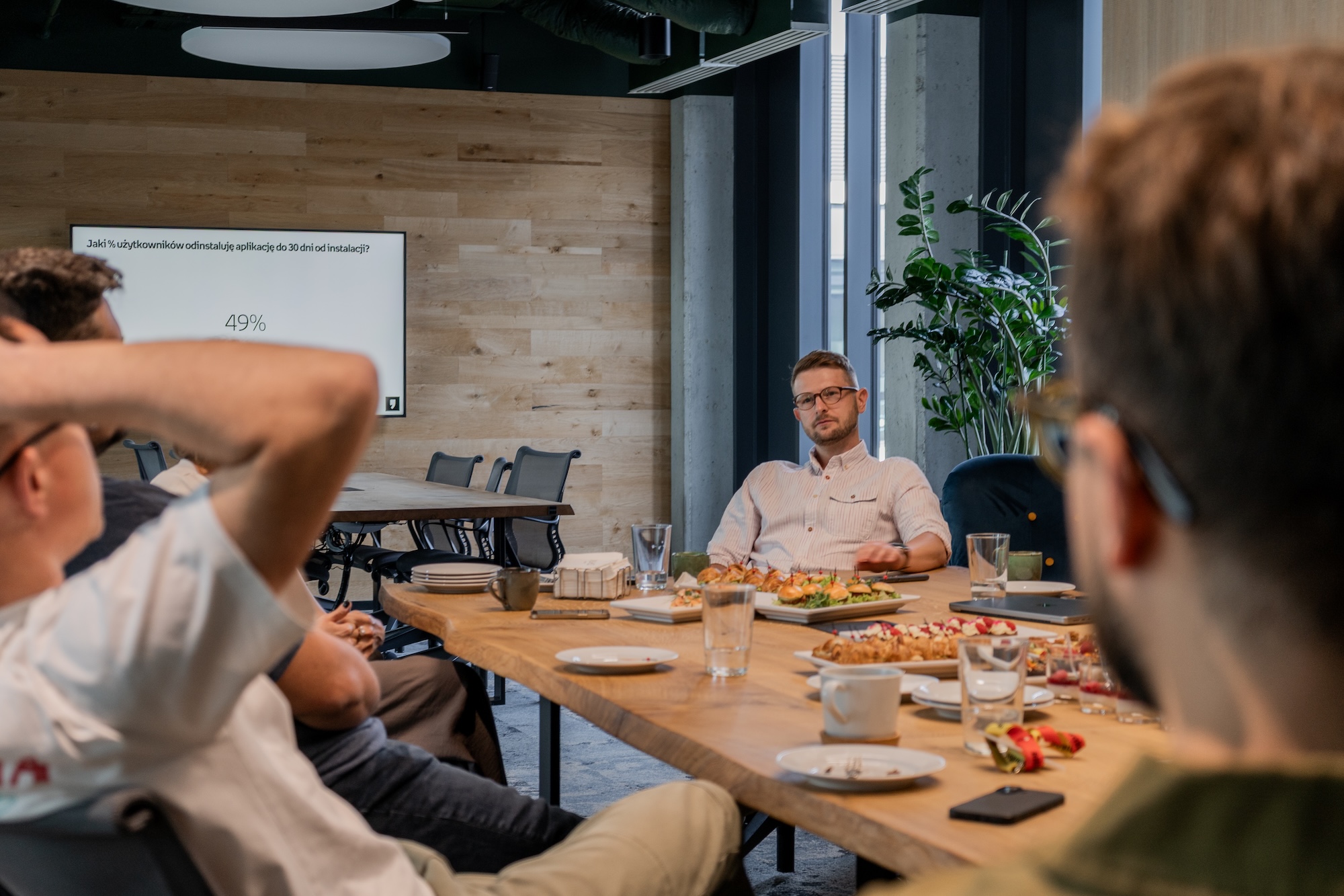 Zdjęcie z wydarzenia Spektrum Offline #4 w SYZYGY Warsaw. Przy stole z poczęstunkiem siedzi Jakub Rak, Head of Mobile w Empik Group, prowadząc rozmowę o lojalności użytkowników. W tle na ekranie widoczny slajd z pytaniem „Jaki % użytkowników odinstaluje aplikację do 30 dni od instalacji?” i wynikiem 49%.